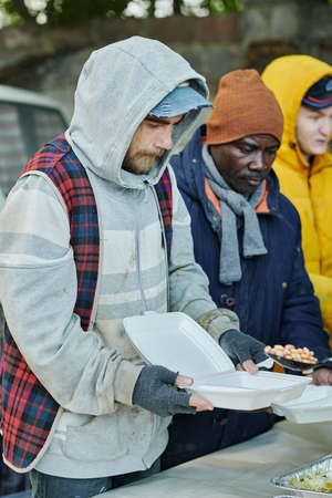 Group of homeless people in warm clothing standing with plastic plates and waiting for food giving by volunteersの写真素材
