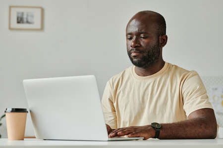 African young businessman sitting at table and typing on laptop, he working online at modern officeの写真素材