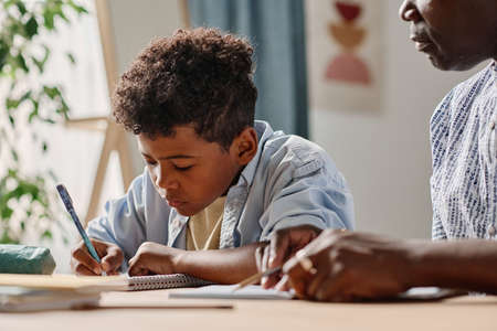 African little boy making notes in notebook while sitting at table together with teacher at homeの写真素材