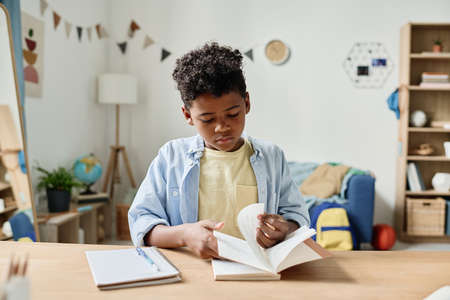 African little boy turning the pages of his book at the table while doing his homework at homeの写真素材