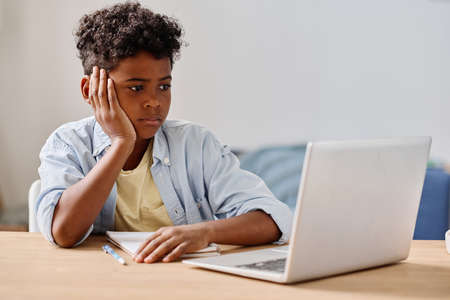 African boy sitting at desk and looking at monitor of laptop during online studying at homeの写真素材