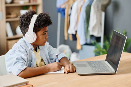 African boy in wireless headphone looking at monitor of laptop and making notes in his notebook during online studyingの写真素材