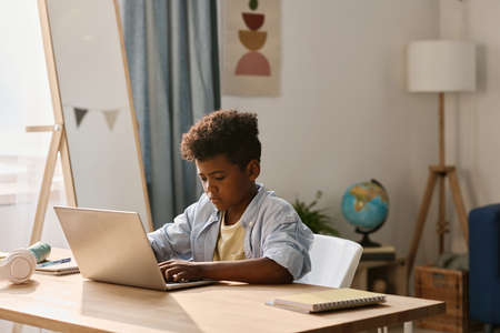 African boy typing on laptop to do his homework online while sitting at desk in his roomの写真素材