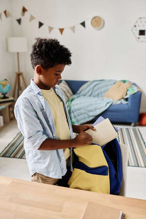 African schoolboy preparing for school packing books in his schoolbag in the roomの写真素材