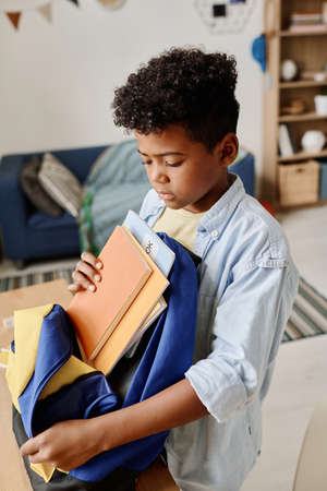 African schoolboy opening his school bag to pack books standing in the room and preparing for school