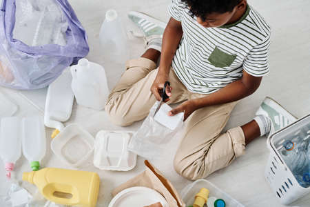 High angle view of African boy sitting on floor and cutting plastic with scissors, he separating rubbish in different containersの写真素材