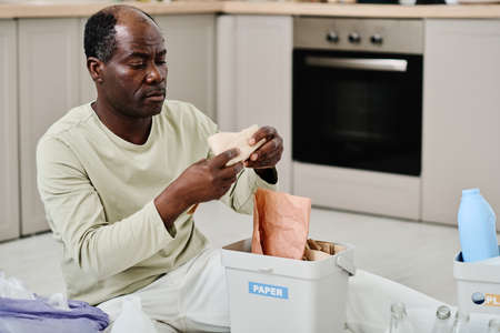 African man putting papers in container, he separating garbage in the kitchen for recyclingの写真素材