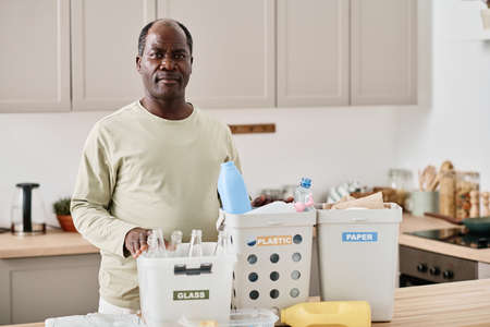 Portrait of African mature man looking at camera while separating plastic, paper and glass in different containers in kitchenの写真素材
