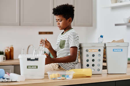 African little boy sorting glass, plastic and paper in different containers while sitting on table in kitchenの写真素材