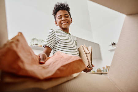 Low angle view of African boy throwing paper in cardboard box for recycling while standing in kitchenの写真素材