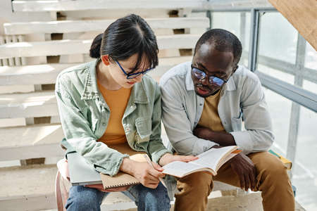 Couple of students preparing for the exams together reading a book while sitting on stairs in school buildingの写真素材