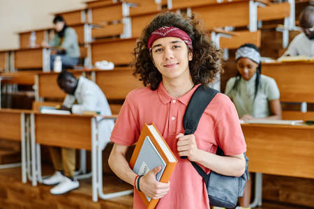 Portrait of student with backpack and book looking at camera while standing at auditorium of university with classmates in backgroundの写真素材