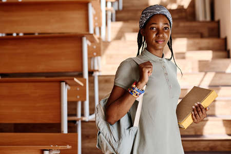 Portrait of African teenage girl with backpack looking at camera standing at empty auditorium of universityの写真素材