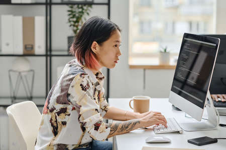 Asian young girl sitting at her workplace in front of computer monitor and typing codes working at officeの写真素材