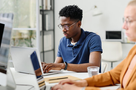African young programmer concentrating on his online work, he typing on laptop while sitting at...