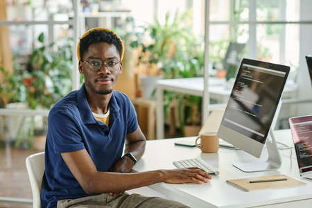 Portrait of African young developer in eyeglasses looking at camera while sitting at his workplace...