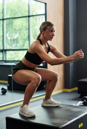 Athletic woman jumping on podium and doing squats during sport training in gymの写真素材
