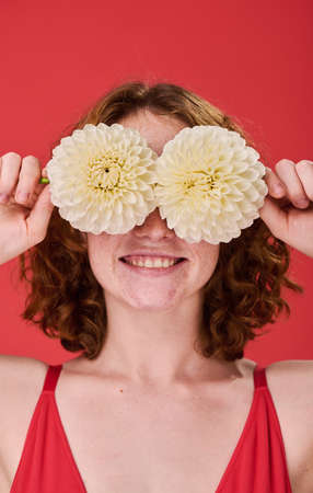 Portrait of young red haired girl holding flowers in front of her eyes and smiling isolated on red backgroundの写真素材
