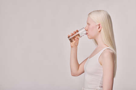 Side view of albino girl with white ong hair drinking water from glass standing on white backgroundの写真素材