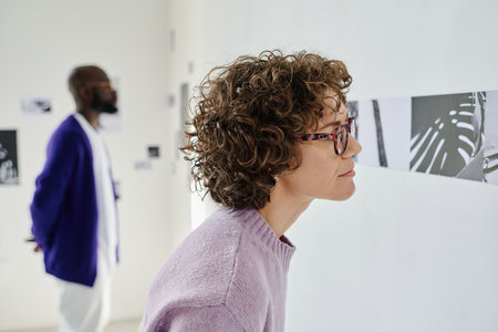 Young woman in eyeglasses examining modern pictures at gallery with man standing in background