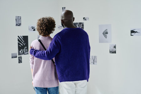 Rear view of multiethnic couple visiting photo gallery, they embracing and watching at photos on the...