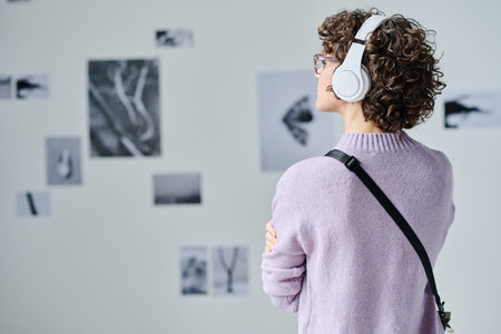 Rear view of young visitor in wireless headphones listening to guide while examining pictures on the...
