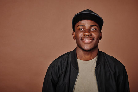 Portrait of African teenage boy in cap smiling at camera isolated on brown background