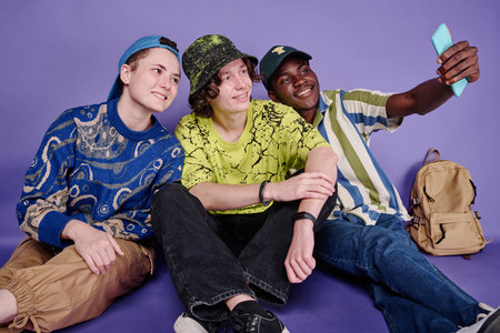 Group of teenagers sitting on floor and making selfie portrait on mobile phone against purple...