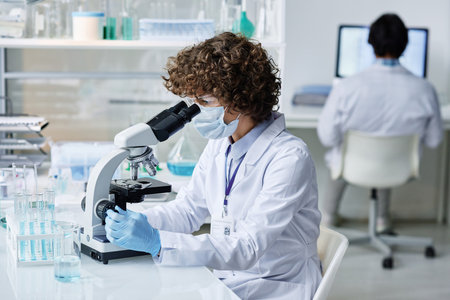 Young woman in labcoat, protective mask and gloves looking in microscope by workplace while carrying out scientific investigationの写真素材