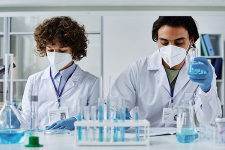 Two young clinicians in labcoats, respirators and gloves working with liquid chemical substances created in laboratory during trialの写真素材
