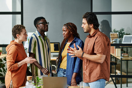 Young multicultural designers having discussion of ideas or working plan while standing in front of each other by workplace with laptopの写真素材