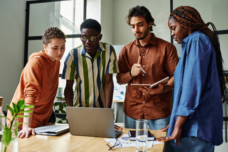 Four young intercultural creative designers looking at graphic data on laptop screen while standing by workplace during slide showの写真素材