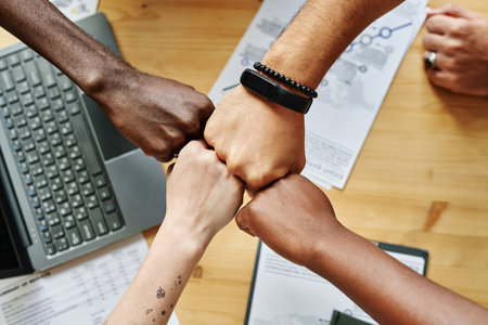 Above angle of four hands of young intercultural employees making punch bump over table with laptop...