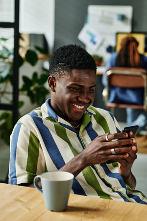 Happy young African American businessman texting in smartphone while sitting by workplace and having coffee at break in officeの写真素材