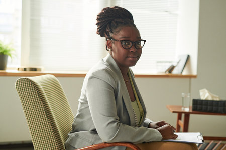 Portrait of African American psychologist in eyeglasses looking at camera while sitting on armchair with document and working at her officeの写真素材