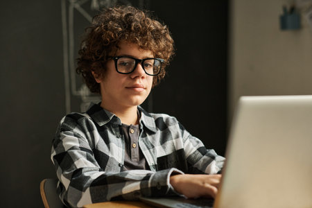 Portrait of schoolboy in eyeglasses looking at camera using laptop sitting at desk during lesson