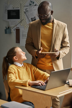 Smiling schoolgirl talking to teacher while using laptop at desk during IT technology lesson