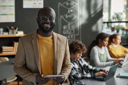Portrait of African American IT teacher smiling at camera standing in classroom with students using...