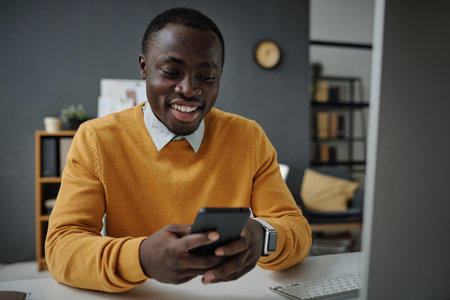 African American businessman typing message on smartphone and smiling while sitting at his workplace at officeの写真素材