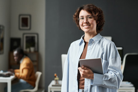 Portrait of young office worker in eyeglasses with tablet pc smiling at camera standing at officeの写真素材