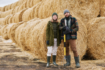 Portrait of young couple of farmers standing outdoors against stack of hay and smiling at camera, they harvesting hay for animalsの写真素材