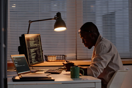 African American IT specialist using his smartphone while working with codes on computer at his...