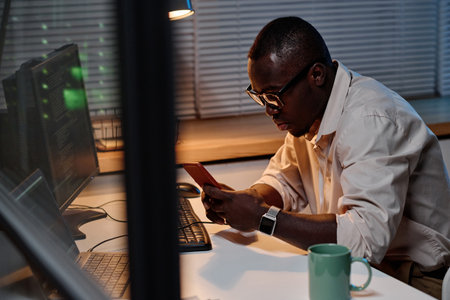 African American IT specialist in eyeglasses working online on smartphone sitting at his workplace...