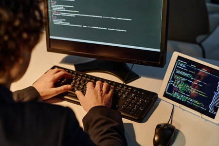 Rear view of young programmer using keyboard to type codes for new software while working in office till late nightの写真素材