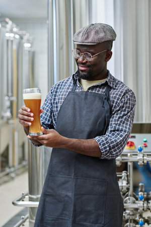 Young African American engineer holding glass of beer in his hands tasting it after brewing