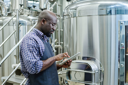 Serious African American engineer working online on digital tablet while standing in workshop of...