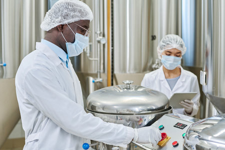Engineer in protective clothing pushing button on equipment while working with assistant at factory