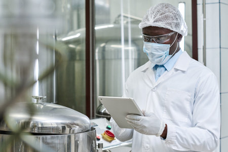 African American engineer in white coat and mask typing on digital tablet while controlling the...