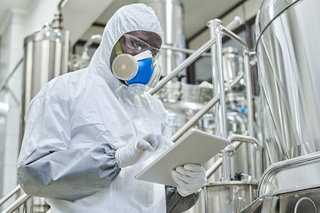 African American chemist in protective costume working online on tablet pc during his work on...