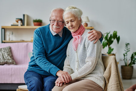 Senior man embracing his wife in depression and supporting her while they sitting on chairs in the roomの写真素材